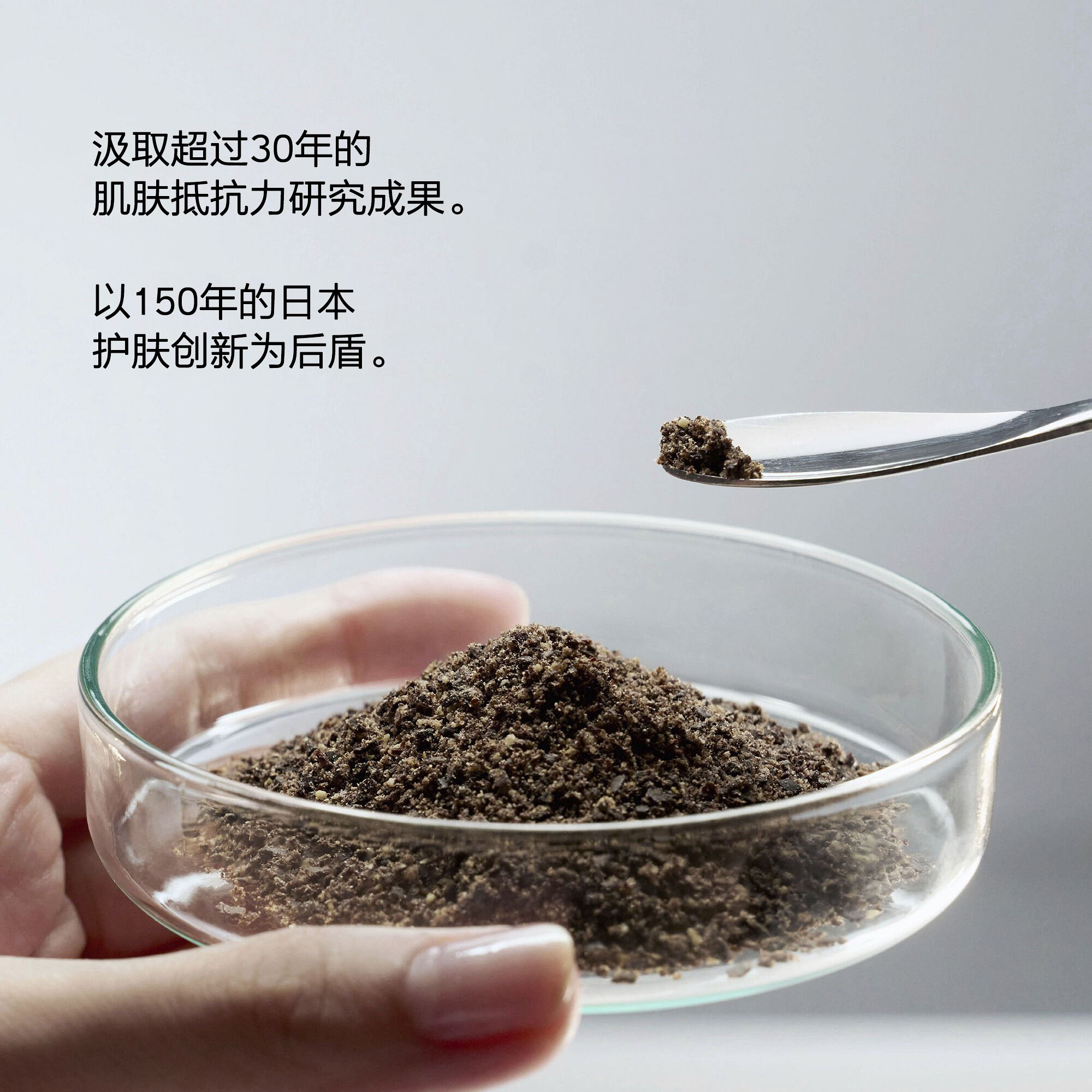文字：‘汲取超过30年的肌肤抵抗力研究成果。以150年的日本护肤创新为后盾。’ 下方，一只手托着装有深色颗粒状粉末的培养皿，上方是小勺舀取少量粉末。